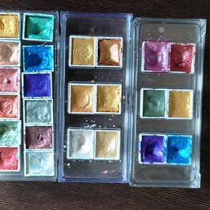 May include: Metallic watercolor paints in a variety of colors, including gold, bronze, blue, and green, are arranged in clear plastic cases. The paints have a shimmering, iridescent finish, perfect for art projects.