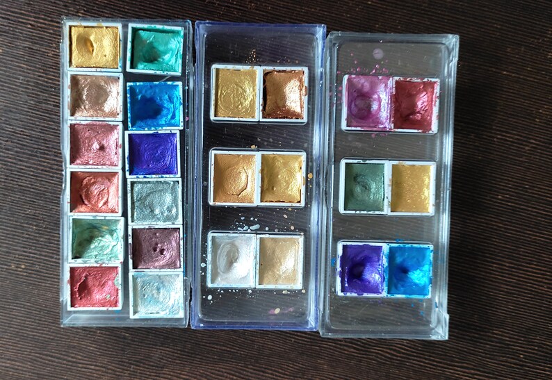 May include: Metallic watercolor paints in a variety of colors, including gold, bronze, blue, and green, are arranged in clear plastic cases. The paints have a shimmering, iridescent finish, perfect for art projects.
