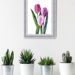 May include: A framed watercolor painting of three tulips in shades of pink and purple, with green leaves. The artwork is in a light blue frame and hangs on a white wall above a shelf with potted plants.