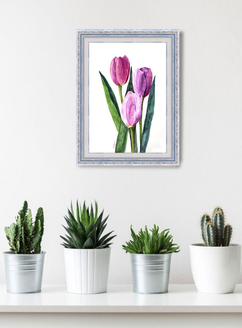 May include: A framed watercolor painting of three tulips in shades of pink and purple, with green leaves. The artwork is in a light blue frame and hangs on a white wall above a shelf with potted plants.