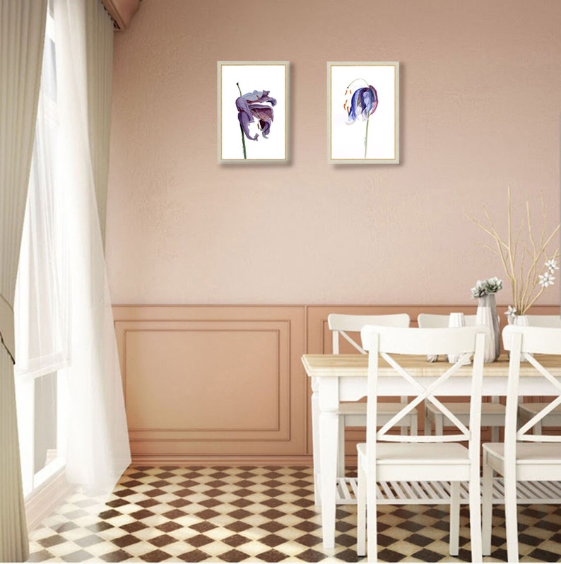 May include: Two framed floral art prints. Each print features a watercolor painting of a purple and blue flower against a white background. The frames are a light wood color. The prints hang on a pink wall above a white table and chairs.