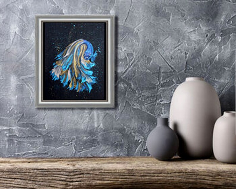 May include: A framed painting of a betta fish with blue and gold brushstrokes against a black background. The artwork is in a silver frame and is displayed on a textured gray wall. Decorative vases are on a wooden surface.