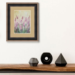 K&ouml;nnte beinhalten: Gerahmtes Kunstwerk mit einem Aquarellgem&auml;lde von Lavendelbl&uuml;ten auf einem Feld. Das Gem&auml;lde hat violette und gr&uuml;ne Farbt&ouml;ne vor einem hellgelben Hintergrund. Der Rahmen ist schwarz mit einem goldenen Innenrand. Schwarze Vasen stehen auf einem Holzregal.