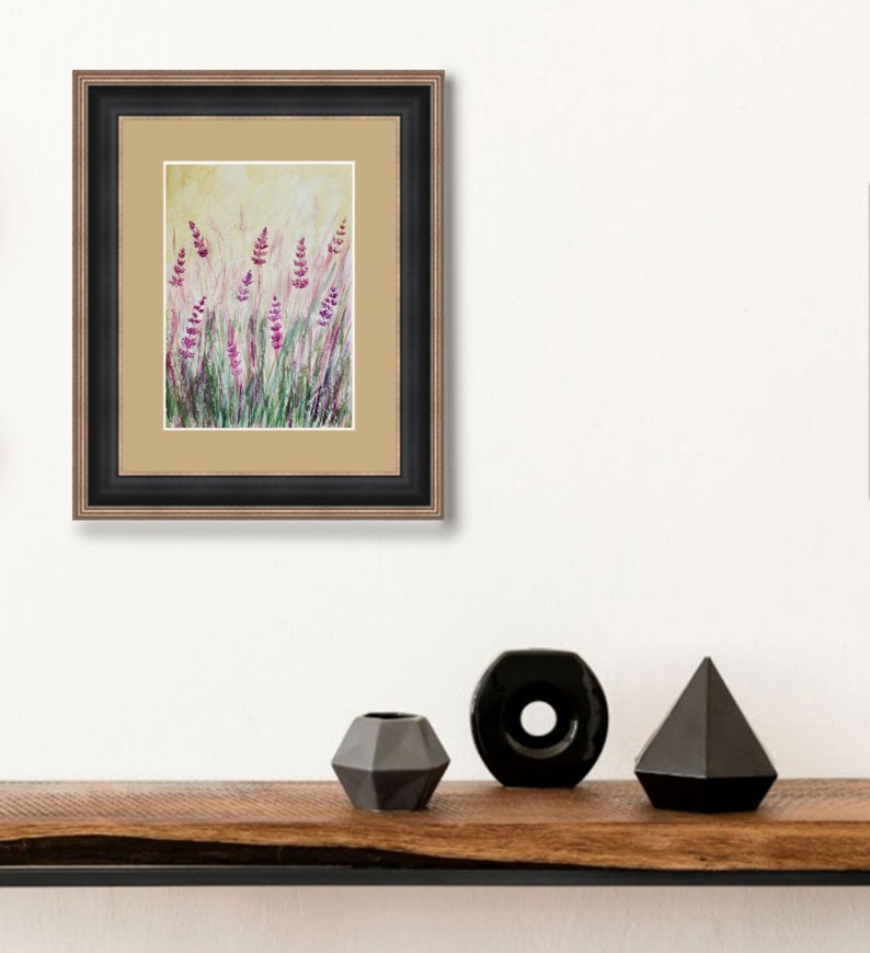 K&ouml;nnte beinhalten: Gerahmtes Kunstwerk mit einem Aquarellgem&auml;lde von Lavendelbl&uuml;ten auf einem Feld. Das Gem&auml;lde hat violette und gr&uuml;ne Farbt&ouml;ne vor einem hellgelben Hintergrund. Der Rahmen ist schwarz mit einem goldenen Innenrand. Schwarze Vasen stehen auf einem Holzregal.