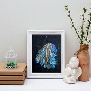May include: A framed abstract painting with blue and gold brushstrokes on a black background. A small white birdcage with a green plant sits on a stack of brown books. A decorative vase with white flowers and a small white angel statue complete the scene.