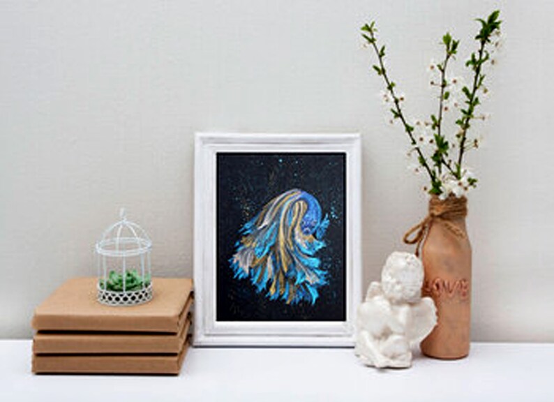 May include: A framed abstract painting with blue and gold brushstrokes on a black background. A small white birdcage with a green plant sits on a stack of brown books. A decorative vase with white flowers and a small white angel statue complete the scene.