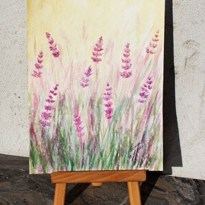 K&ouml;nnte beinhalten: Aquarellmalerei von Lavendelbl&uuml;ten auf einem Feld. Das Kunstwerk zeigt lila Lavendelbl&uuml;ten und gr&uuml;nes Gras vor einem sanften gelben Hintergrund. Das Gem&auml;lde wird auf einer kleinen Holzstaffelei ausgestellt.