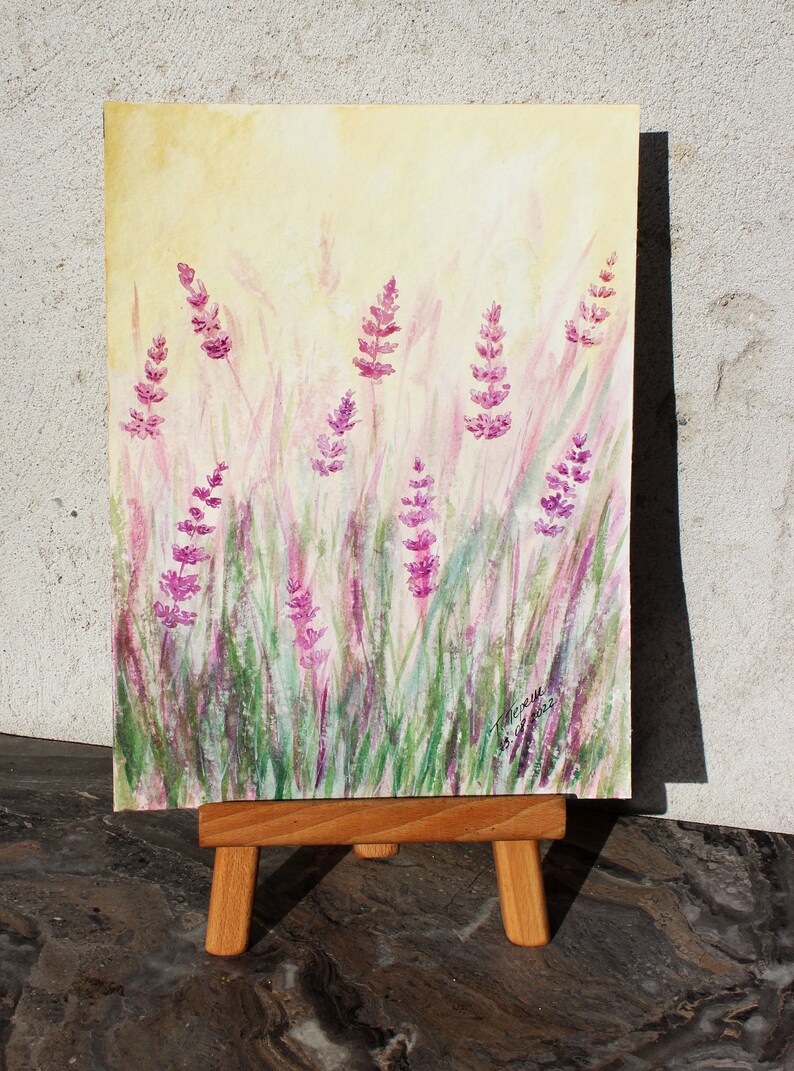 K&ouml;nnte beinhalten: Aquarellmalerei von Lavendelbl&uuml;ten auf einem Feld. Das Kunstwerk zeigt lila Lavendelbl&uuml;ten und gr&uuml;nes Gras vor einem sanften gelben Hintergrund. Das Gem&auml;lde wird auf einer kleinen Holzstaffelei ausgestellt.