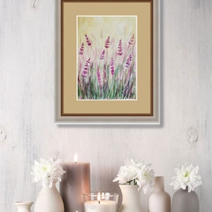 K&ouml;nnte beinhalten: Gerahmtes Aquarellgem&auml;lde mit Lavendelbl&uuml;ten in einem Feld. Das Kunstwerk zeigt lila und gr&uuml;ne Farbt&ouml;ne vor einem hellgelben Hintergrund. Das Gem&auml;lde wird &uuml;ber einem Holzregal mit Kerzen und wei&szlig;en Blumenarrangements ausgestellt.