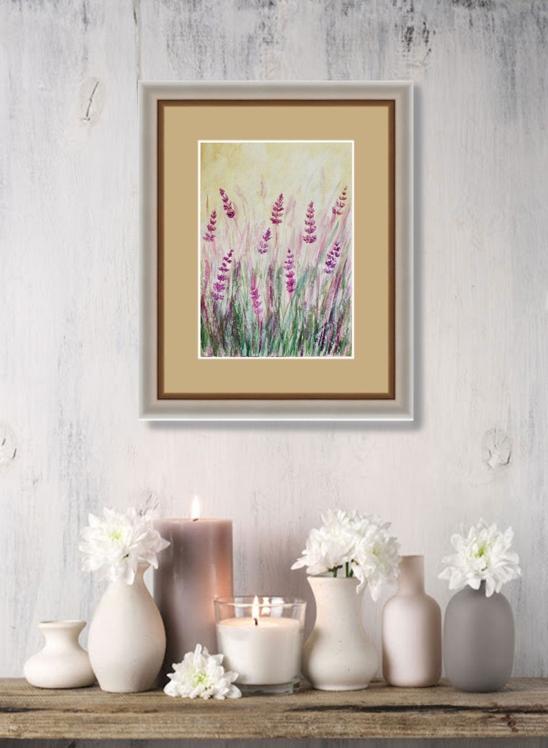 K&ouml;nnte beinhalten: Gerahmtes Aquarellgem&auml;lde mit Lavendelbl&uuml;ten in einem Feld. Das Kunstwerk zeigt lila und gr&uuml;ne Farbt&ouml;ne vor einem hellgelben Hintergrund. Das Gem&auml;lde wird &uuml;ber einem Holzregal mit Kerzen und wei&szlig;en Blumenarrangements ausgestellt.