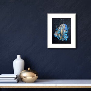 May include: A framed abstract painting with a white border. The artwork features a blue and gold design on a black background. A white vase, a gold decorative sphere, and a stack of books are on a white table.