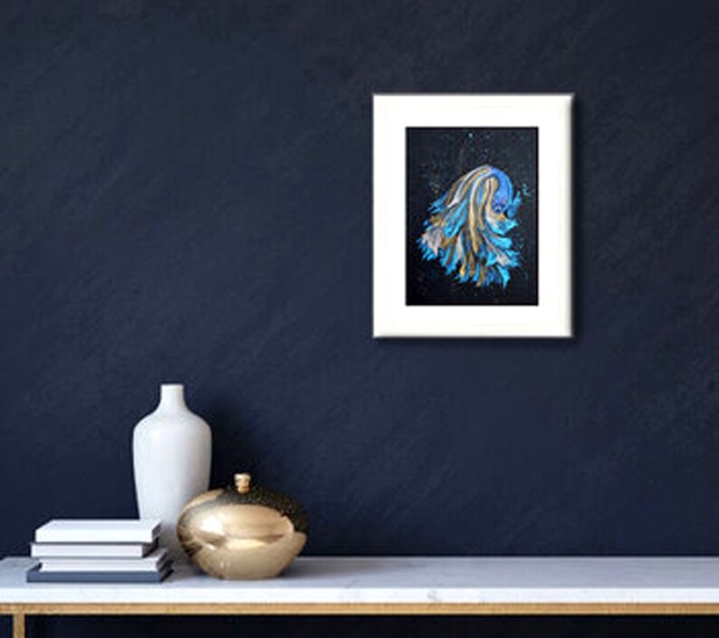 May include: A framed abstract painting with a white border. The artwork features a blue and gold design on a black background. A white vase, a gold decorative sphere, and a stack of books are on a white table.