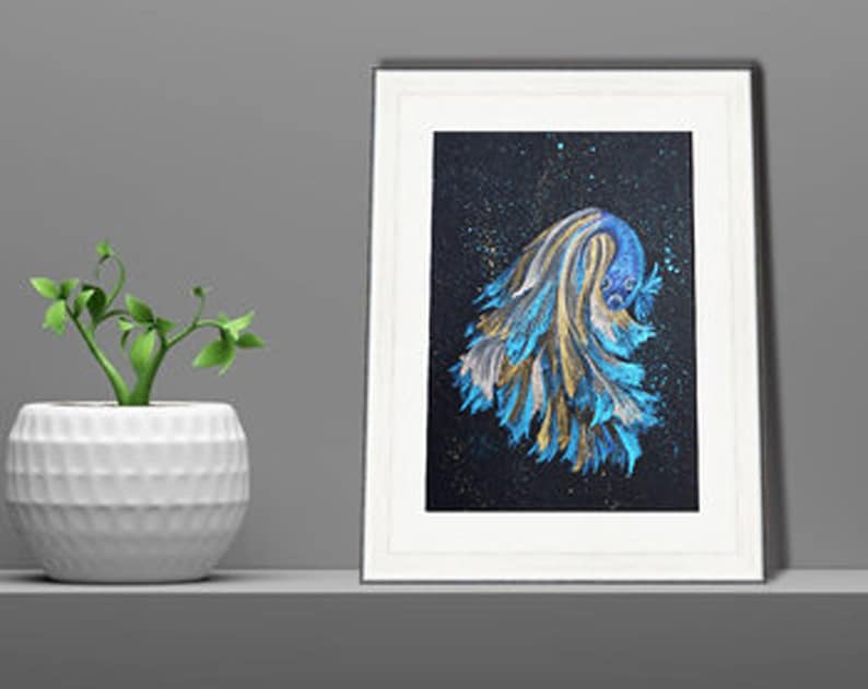 May include: Framed artwork featuring a vibrant betta fish illustration. The fish is depicted with flowing blue, gold, and white fins against a dark, speckled background. The artwork is in a white frame, displayed on a shelf with a small potted plant.