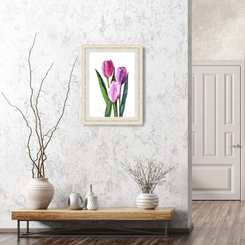 May include: A framed watercolor painting of three tulips in shades of purple and pink, with green leaves. The artwork is displayed on a white background within a cream-colored frame. The painting is hung on a textured, light gray wall.