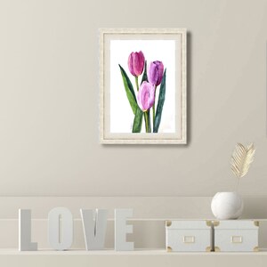 May include: A framed watercolor painting of three tulips in shades of pink and purple, with green leaves. The artwork is in a white ornate frame. The painting is displayed on a neutral-colored wall.