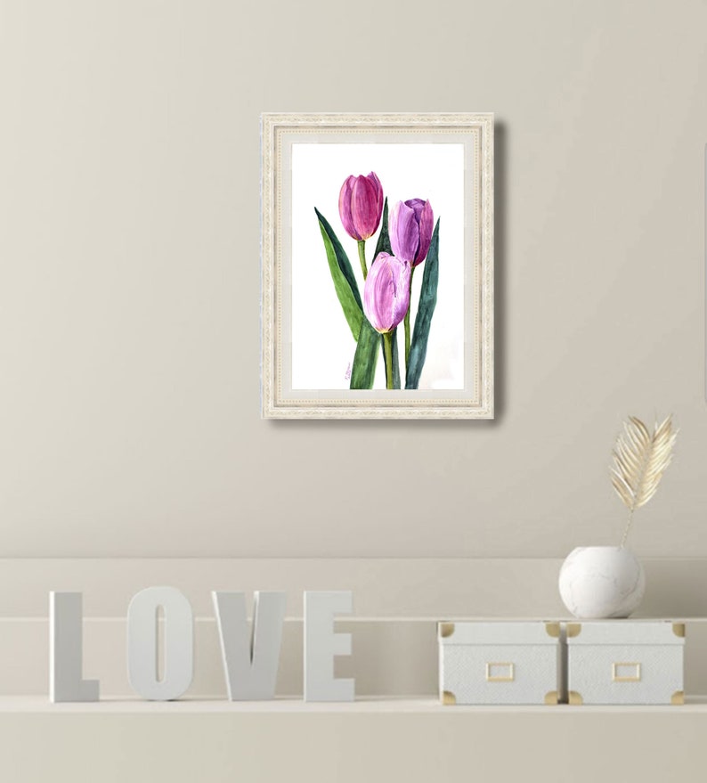 May include: A framed watercolor painting of three tulips in shades of pink and purple, with green leaves. The artwork is in a white ornate frame. The painting is displayed on a neutral-colored wall.