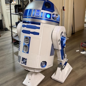 Könnte beinhalten: Eine lebensgroße R2-D2-Droiden-Nachbildung mit einem weißen zylindrischen Körper und blauen sowie silbernen Akzenten. Die Kuppel ist silberfarben mit blauen Paneelen und einem leuchtend blauen Licht. Er steht auf zwei seitlichen Beinen mit flachen Füßen.