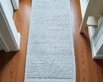 May include: A light gray rectangular bath mat with a textured, nubby surface. The mat is placed on a hardwood floor, and is surrounded by a white door frame, a small potted plant, and a white cabinet.
