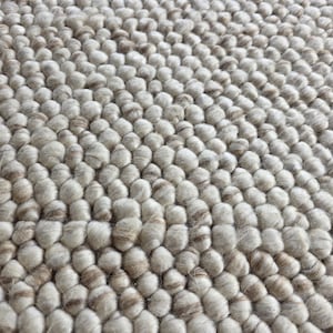 Puede incluir: Primer plano de una alfombra texturizada de tonos neutros. La superficie de la alfombra está cubierta de pequeños elementos redondeados y elevados, creando una apariencia rugosa y táctil. La paleta de colores es principalmente blanquecina con sutiles acentos marrones.