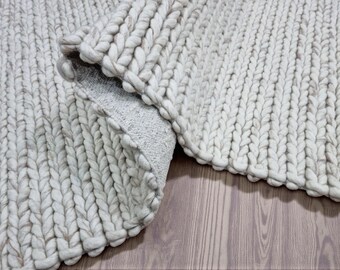 May include: A rolled-up, chunky knit rug in a light gray color. The rug is made of thick, braided yarn, creating a textured surface. The underside of the rug is a lighter gray fabric. The rug is partially rolled on a wooden floor.