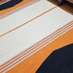 May include: A rectangular woven rug featuring a color-blocked design with large sections of cream, vibrant orange, and dark navy blue. The rug is accented by thin, light-colored horizontal stripes and includes a white fringe along the short edge.