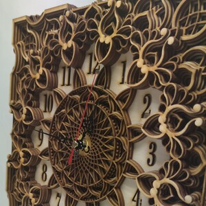 Könnte beinhalten: Quadratische Wanduhr aus Holz mit einem mehrschichtigen Blumendesign. Das Zifferblatt hat ein zentrales Mandala-Muster und schwarze Zeiger mit einem roten Sekundenzeiger. Die Zahlen sind aus dem Holz geschnitten.