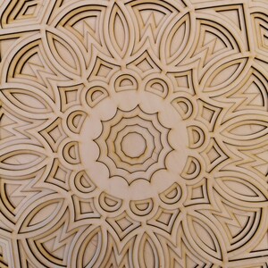May include: A detailed, laser-cut wooden mandala with a layered design. The natural wood tones are accented by dark brown outlines, creating a striking contrast. The intricate pattern features geometric shapes and floral elements, perfect for home decor.