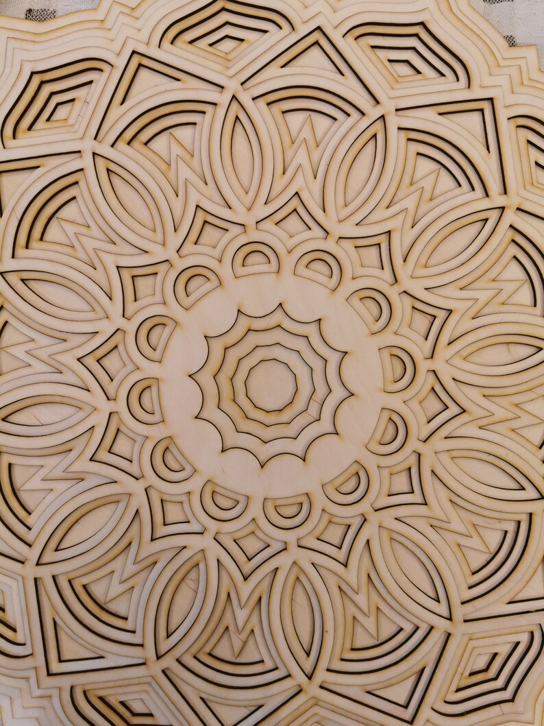 May include: A detailed, laser-cut wooden mandala with a layered design. The natural wood tones are accented by dark brown outlines, creating a striking contrast. The intricate pattern features geometric shapes and floral elements, perfect for home decor.