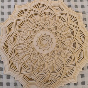 May include: A layered wooden mandala with a detailed, symmetrical design. The natural wood tones and intricate cut-out patterns create a decorative, artistic piece. The mandala features geometric shapes and floral-like elements.