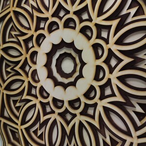 May include: A layered wooden mandala with a complex geometric design. The artwork features alternating layers of light and dark brown wood, creating a three-dimensional effect. The design is circular, with intricate patterns.