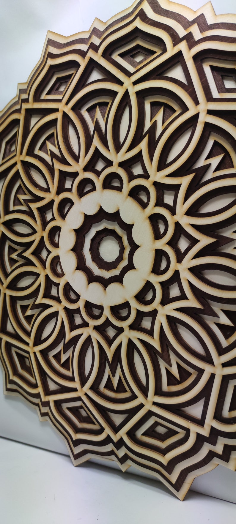 May include: A layered wooden mandala with a complex geometric design. The artwork features alternating layers of light and dark brown wood, creating a three-dimensional effect. The design is circular, with intricate patterns.