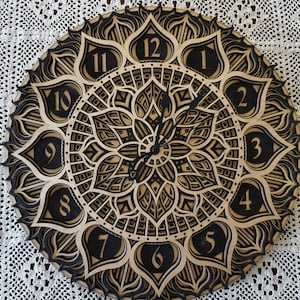 Könnte beinhalten: Eine runde, mehrschichtige Holzuhr mit einem Mandala-Design. Die Uhr hat eine natürliche Holzfarbe mit schwarzen Akzenten, einschließlich der Zahlen und der komplizierten Muster. Die Zeiger der Uhr sind schwarz.