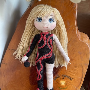 May include: Crocheted doll with long blonde hair, wearing a black and red outfit. The doll is sitting on a wooden surface.