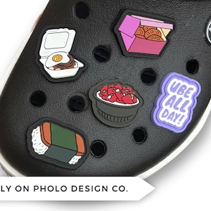 May include: A set of six colorful shoe charms featuring various food items, including a bowl of ube, a plate of spam musubi, a container of fried chicken, a plate of loco moco, and a purple charm with the text "UBE ALL DAY!"