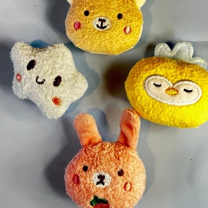 May include: Four plush toy ornaments in the shape of a bear, star, owl, and bunny. The bear is yellow with a blue and white striped hat. The star is white. The owl is yellow with closed eyes. The bunny is peach with a strawberry.