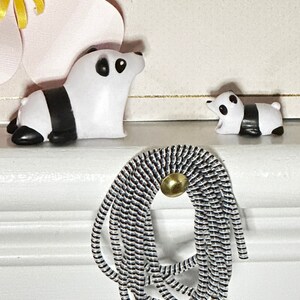 May include: Two panda figurines, one large and one small, are displayed on a white surface. The pandas are white with black markings. A decorative element made of black and white cord with a gold-colored button is also present.