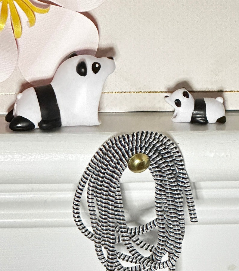 May include: Two panda figurines, one large and one small, are displayed on a white surface. The pandas are white with black markings. A decorative element made of black and white cord with a gold-colored button is also present.