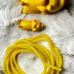 May include: A bright yellow lion-shaped toy and a smaller version of the same toy are lying on a white, fluffy surface. A coiled yellow cable protector is also in the image. The toys have black eyes and a small tail.