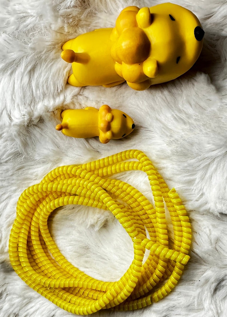 May include: A bright yellow lion-shaped toy and a smaller version of the same toy are lying on a white, fluffy surface. A coiled yellow cable protector is also in the image. The toys have black eyes and a small tail.