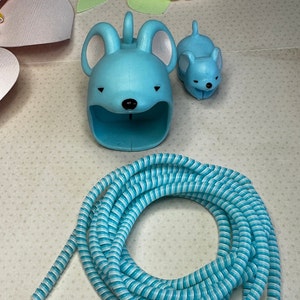 May include: A light blue cable organizer shaped like a mouse, a smaller mouse figurine, and a coiled blue and white striped cable. The mouse-shaped organizer has large ears and a slot for holding cables. A strand of faux pearls is also visible.
