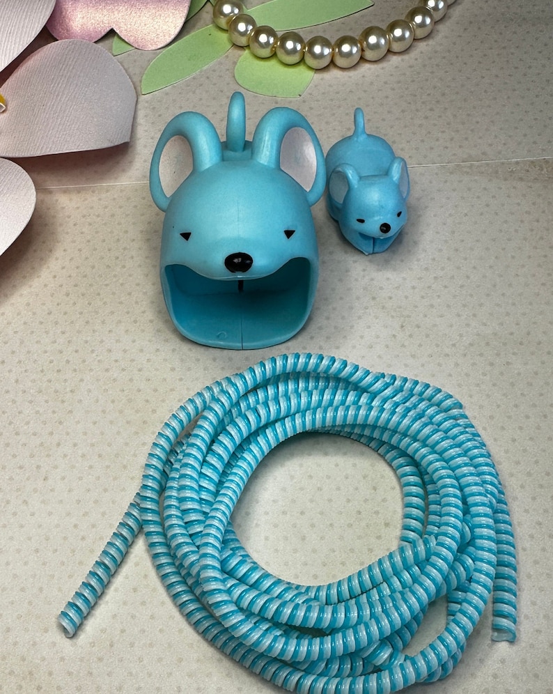 May include: A light blue cable organizer shaped like a mouse, a smaller mouse figurine, and a coiled blue and white striped cable. The mouse-shaped organizer has large ears and a slot for holding cables. A strand of faux pearls is also visible.
