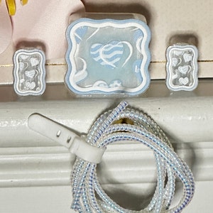May include: A set of clear, heart-themed resin charms and a beaded lanyard. The charms feature a light blue heart design on a white background. The lanyard is made of iridescent beads, held together with a white plastic clasp.