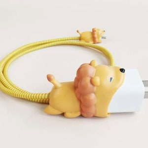 May include: A yellow and brown lion-shaped phone charger with a matching cable. The charger has a white plug and a small lion figure on the cable. The lion figures have a light brown mane and tail.