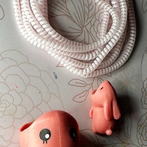 May include: A coiled white cable with a spiral design is positioned above two pink figurines. One figurine is an octopus with black eyes, and the other is a bunny. The background has a floral pattern.