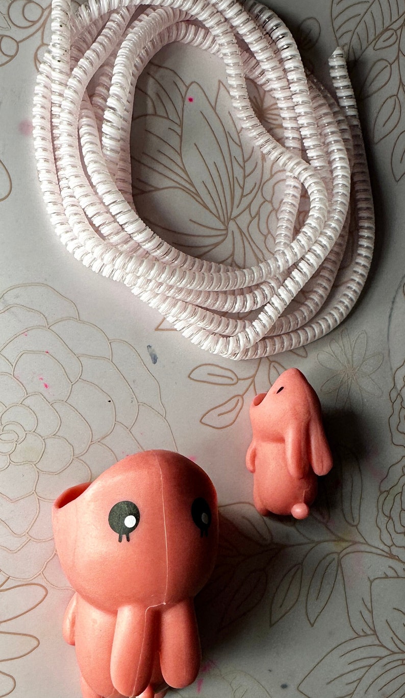 May include: A coiled white cable with a spiral design is positioned above two pink figurines. One figurine is an octopus with black eyes, and the other is a bunny. The background has a floral pattern.