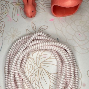 May include: An overhead shot features a coiled, pink and white cable, along with two coral-colored bunny-shaped items. One bunny is a small figurine, and the other is a larger, open-mouthed item. The items are on a floral patterned surface.