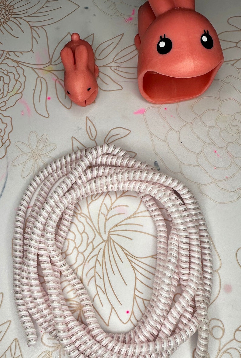 May include: An overhead shot features a coiled, pink and white cable, along with two coral-colored bunny-shaped items. One bunny is a small figurine, and the other is a larger, open-mouthed item. The items are on a floral patterned surface.