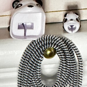 May include: A white panda-shaped accessory with black accents, featuring a backpack design and a smaller panda head. A black and silver cord with a gold-colored bead is in the foreground. The items are on a white surface.