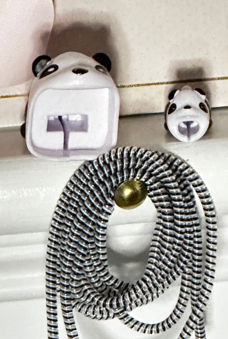 May include: A white panda-shaped accessory with black accents, featuring a backpack design and a smaller panda head. A black and silver cord with a gold-colored bead is in the foreground. The items are on a white surface.