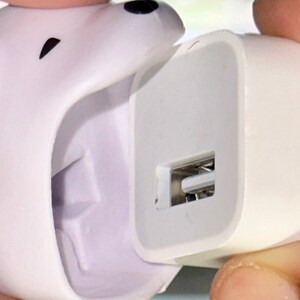 May include: A white USB charger with a polar bear design. The charger has a USB port and is being held by a person. The polar bear's head is on the left side of the charger.